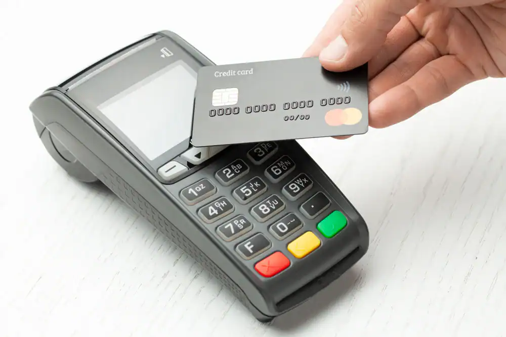 A hand holds a credit card near a payment terminal, demonstrating merchant processing in Anne Arundel County, MD. The contactless transaction occurs on a white surface, with the terminal displaying its keypad and colored buttons.