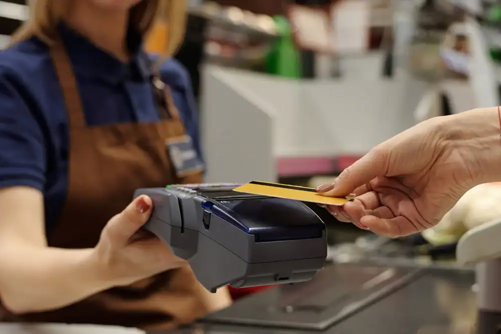 A cashier in a brown apron holds a payment terminal while a customer in Anne Arundel County, MD, extends a yellow credit card to make a contactless merchant processing payment at the checkout counter.