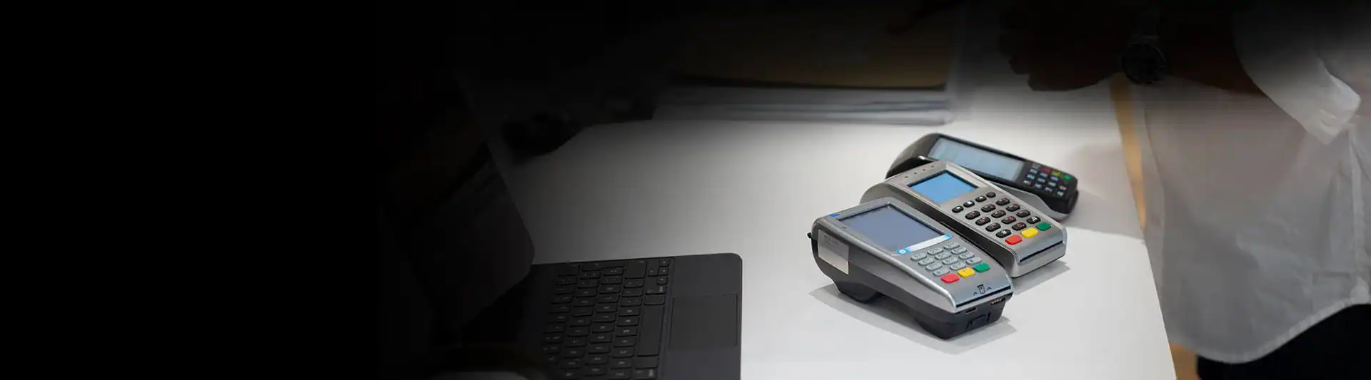 Two point-of-sale card payment terminals are on a white desk next to a laptop in an MD office, highlighting merchant processing in Anne Arundel County, with a person partially visible on the right side of the image.