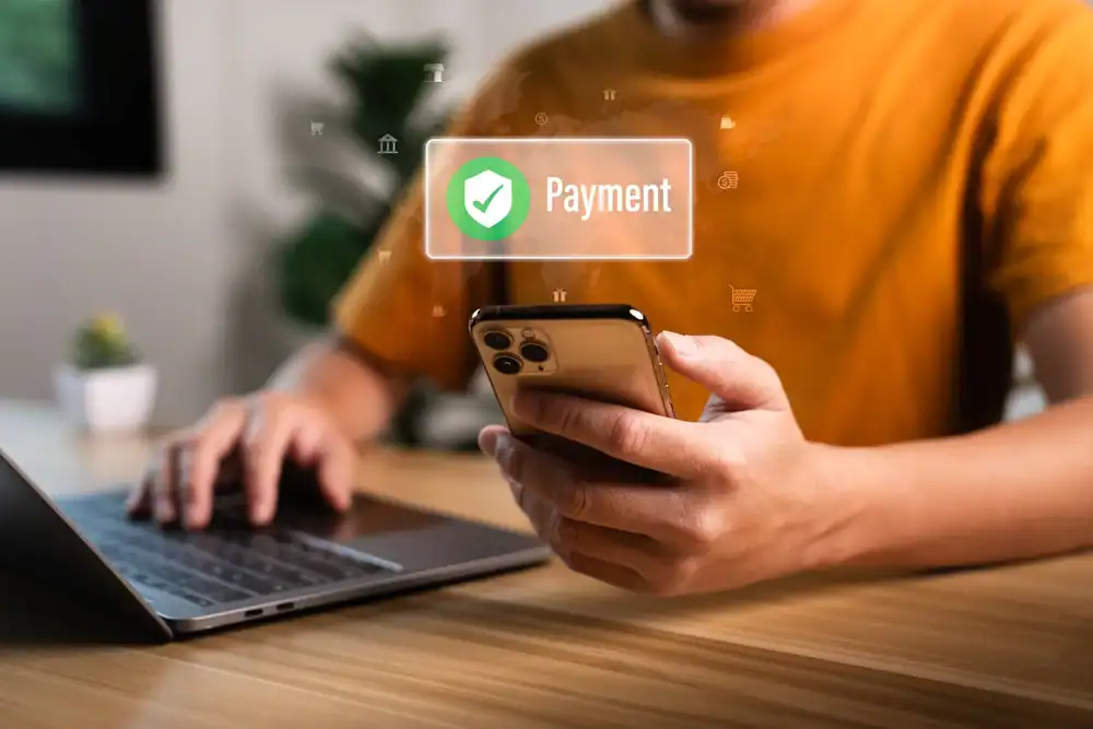 A person in an orange shirt uses a smartphone and laptop at a wooden table, with a holographic "Payment" icon and checkmark above the phone—highlighting successful online merchant processing in Anne Arundel County, MD.