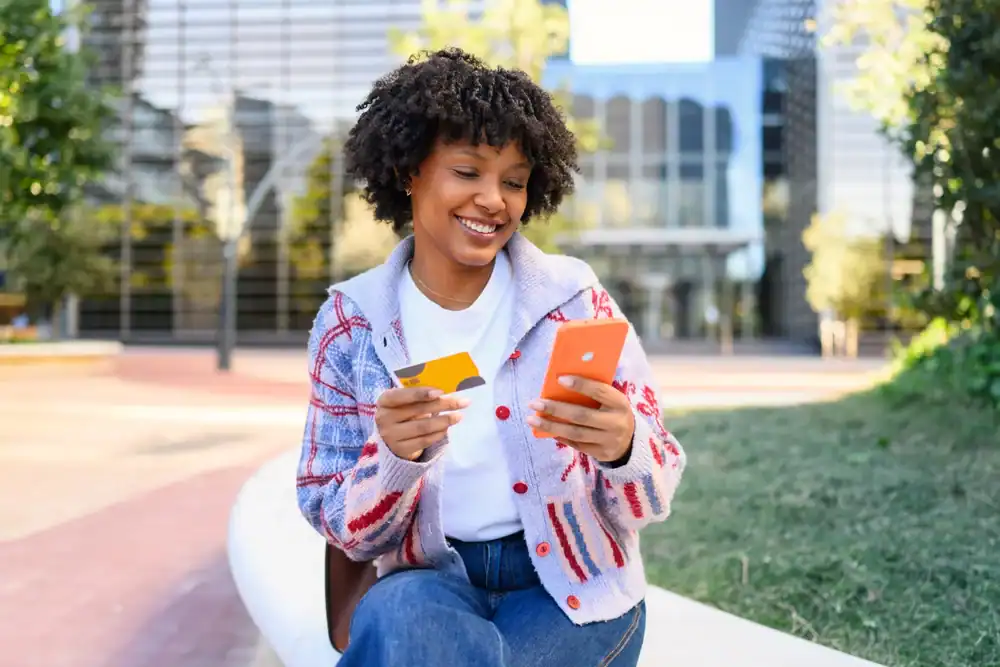 Young woman smiling while making mobile payment with credit card outdoors in city park.