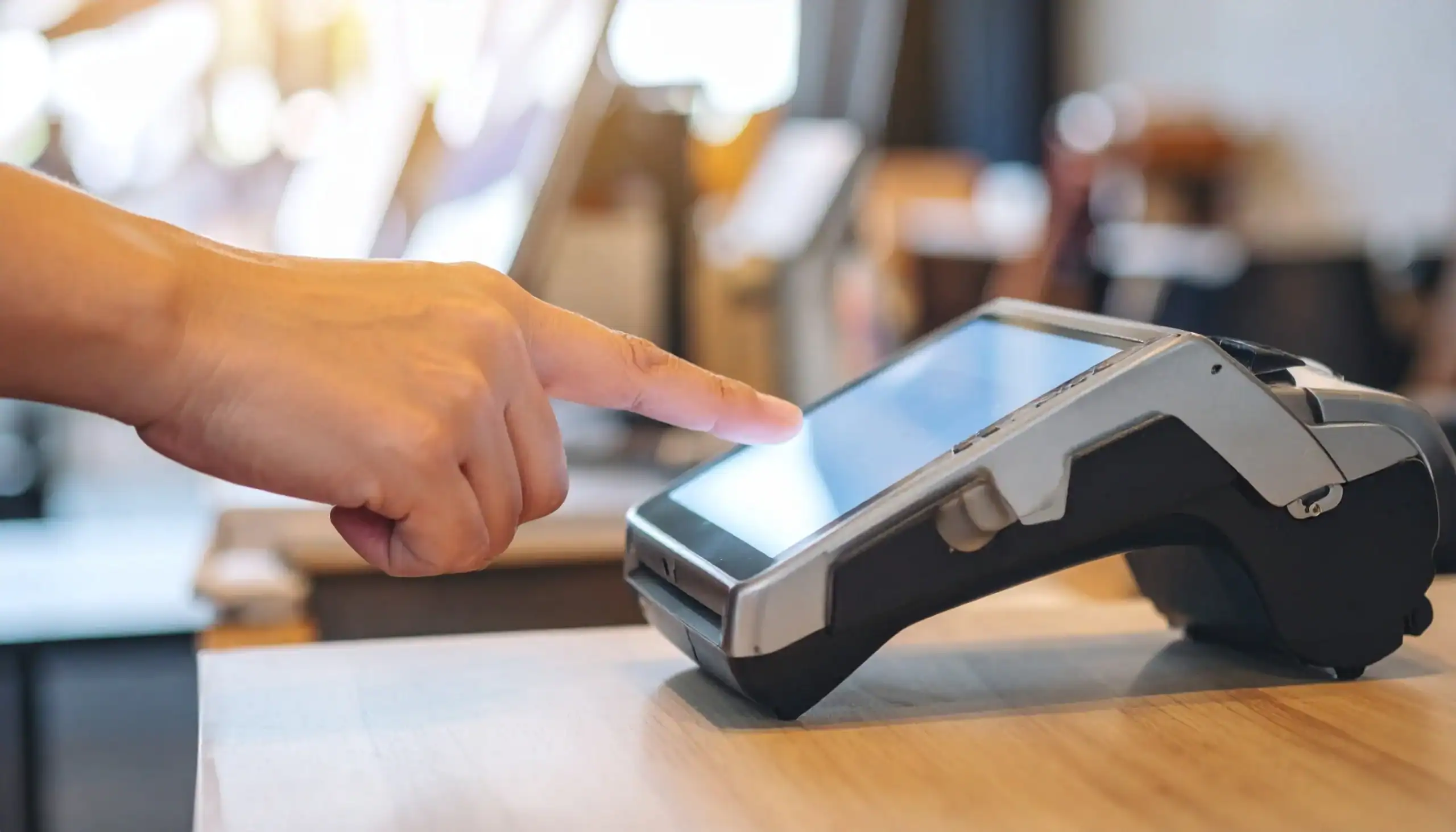 Close-up of a person's hand using a touchscreen POS payment device on a wooden surface.